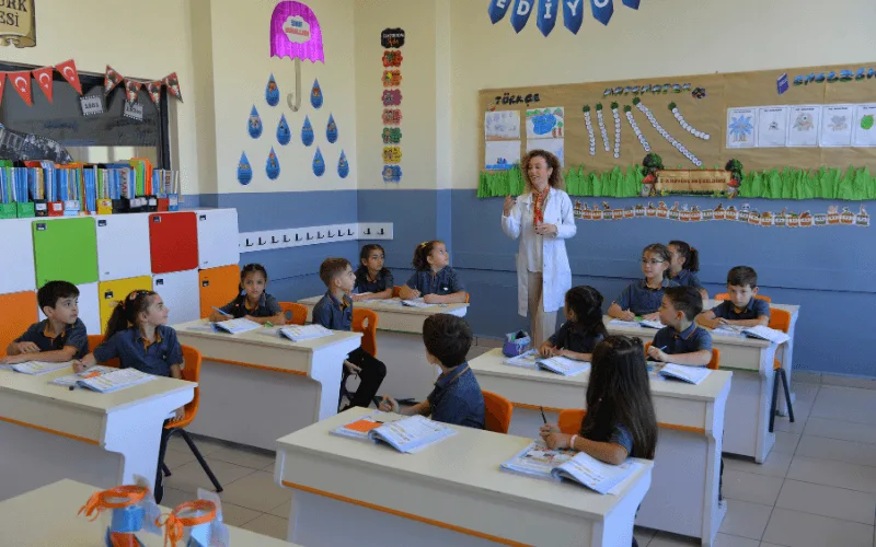 İlkokul sınıfında öğretmen tahtada ders anlatırken öğrenciler sırada dinliyor